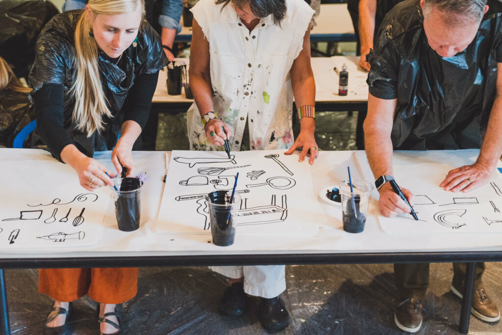 Three individuals painting line art in black paint on white butcher paper.